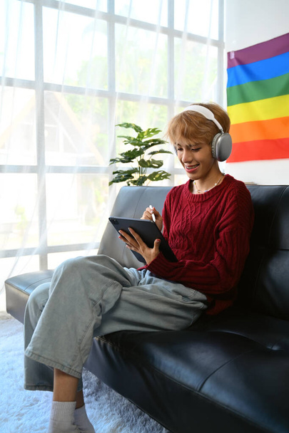 Atractivo joven gay hombre con auriculares usando tableta digital, navegar por las redes sociales, ver películas mientras se relaja en el sofá en casa. - Foto, imagen
