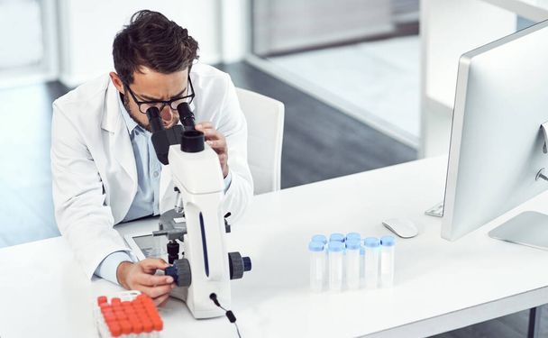 Ничто не может его беспокоить, пока он работает. сфокусированный молодой ученый-мужчина, смотрящий в микроскоп в лаборатории - Фото, изображение
