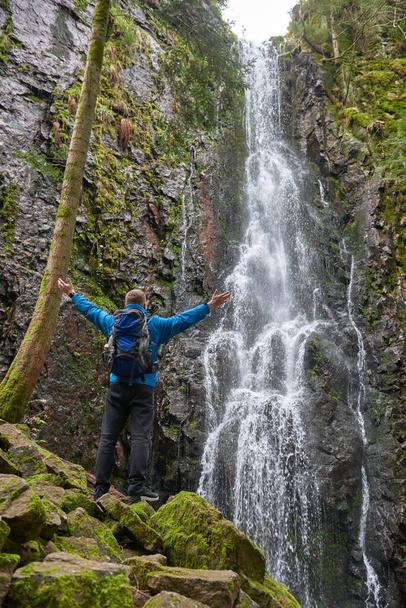 Tourist attraction of Germany - falls of Burgbach Waterfall near Schapbach, Black Forest, Baden-Wurttemberg, Germany. Man hiker in blue jacket standing on stone and looks at flow of falling water - Fotografie, Obrázek
