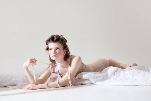 Beautiful, tender young girl with pink blusher on cheeks lying in retro lingerie on blanket against grey studio background. Sensuality. Concept of female beauty, art, fashion, emotions. Pin up style - Valokuva, kuva