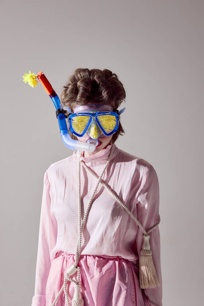 Portrait of young girl in pink outfit posing in water diving mask against grey studio background. Uniqueness and weirdness. Concept of female beauty, art, femininity, fashion, emotions - Photo, Image