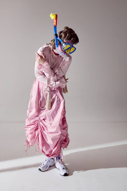 Portrait of young girl in pink outfit posing in water diving mask against grey studio background. Uniqueness and extraordinary look. Concept of female beauty, art, femininity, fashion, emotions - Photo, image
