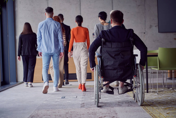 A diverse group of businessmen, including a businessman in wheelchair, confidently stride together through a modern, spacious office, epitomizing collaboration, inclusivity, and strength in unity.  - Valokuva, kuva