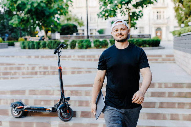 Красивый мужчина держит ноутбук в руке, стоит рядом с электрическим скутером. Активный образ жизни, турист путешественник. Экология и чистый экологический вид транспорта - Фото, изображение