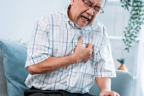 An agonizing senior man suffering from chest pain or heart attack alone in his living room. Serious health problem and feeling unwell concept for seniors. - Photo, image