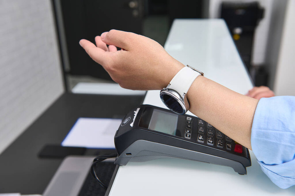 Κοντινό χέρι μιας γυναίκας που πληρώνει λογαριασμό με έξυπνο ρολόι με τεχνολογία NFC. Πελάτης που κάνει ανέπαφη πληρωμή χρησιμοποιώντας SmartWatch. Επικοινωνία κοντά στο πεδίο. Ανθρώπους. Εξυπηρέτηση πελατών. Ασύρματες τεχνολογίες - Φωτογραφία, εικόνα