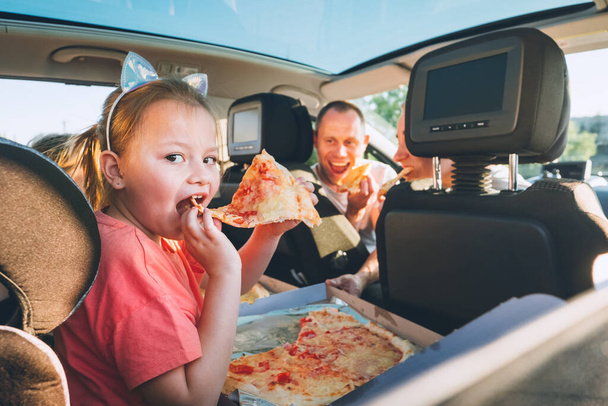 Little girl with open mouth portrait eating Italian pizza sitting in modern car with mother and father. Happy family moments, childhood, fast food eating or auto journey lunch break concept image.	 - Photo, Image