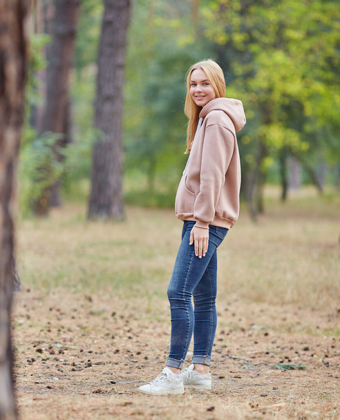 Attractive blue eyed blonde woman walk on the city park. Girl wear beige hoodie, pink bag and look happy and smiles. Portrait of a joyful young woman enjoying in autumn park. Relax in nature. - Photo, Image