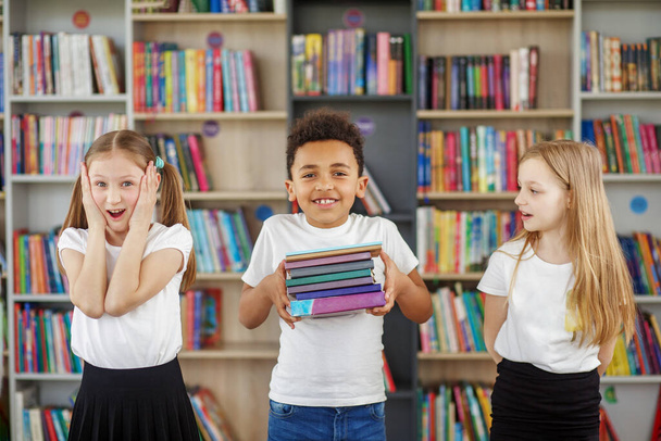 Niños sosteniendo una pila de libros en la biblioteca. Compañeros multiétnicos divirtiéndose. Día Mundial del Libro. Beneficios de la lectura cotidiana. - Foto, imagen