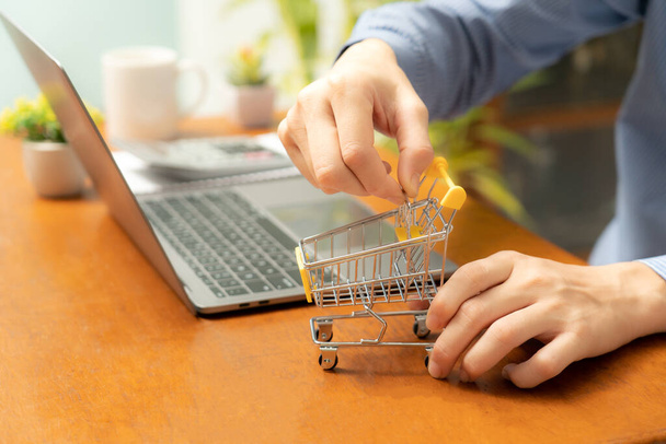 The concept of online shopping materializes as a shopping cart, accompanied by a laptop, is positioned on the table,and a person's hand firmly grasps the cart.online shopping concept shopping.close up - Photo, image