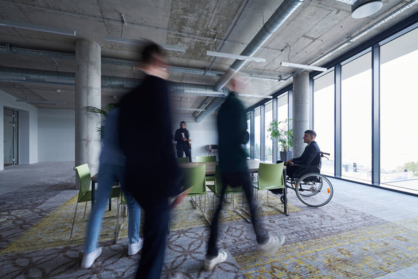 A businessman in a wheelchair sits at a table in a large, modern office, while his colleagues gather around, their steps blurred, symbolizing inclusivity, support, and unity in the face of challenges - 写真・画像