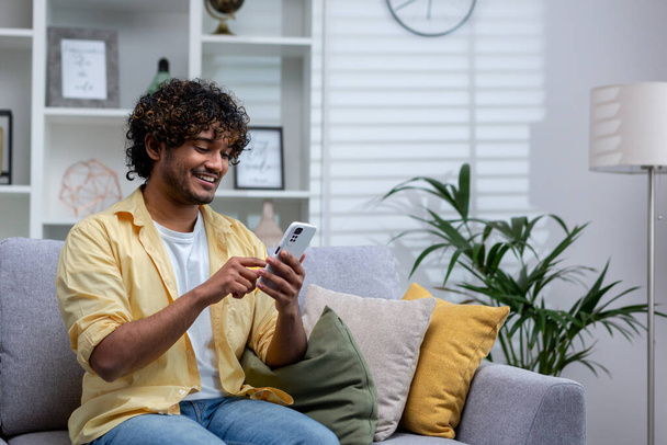 Un joven hispano está sentado en casa en el sofá de la sala de estar, el hombre sostiene un teléfono en sus manos, usando una aplicación en línea en un teléfono inteligente, navegando por Internet y escribiendo mensajes. - Foto, Imagen