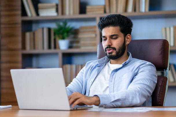 Junge indische Geschäftsmann ceo executive sitzt am Schreibtisch mit Laptop in zeitgenössischen Büro. Seriöse Finanzanalysten, die mit Online-Daten arbeiten. Geschäftstechnologisches Konzept. - Foto, Bild