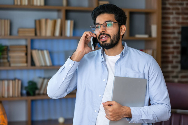 Een lachende zakenman gekleed in een blauw shirt, pratend op zijn telefoon met laptop op kantoor. concept van werken op afstand en freelance werk. jong modern indisch succesvol man in bril - Foto, afbeelding