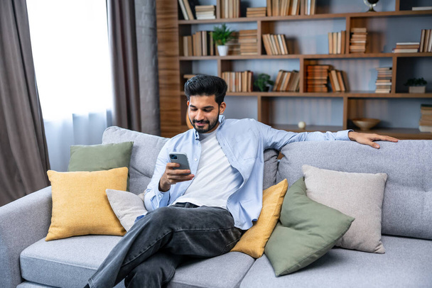 indian millennial bearded man using smart phone for scrolling social media at home - Fotó, kép