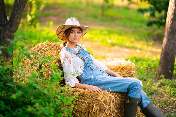 Belle cow-girl blonde en chapeau de paille, chemise à carreaux et denim assis sur un fond vert nature. Oeuvre d'art colorée. - Photo, image