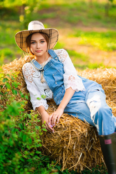 Beautiful blonde cowgirl in straw hat, checkered shirt and denim overall sitting on a straw on green nature background. Colorful art work. - 写真・画像