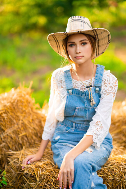 Beautiful blonde cowgirl in straw hat, checkered shirt and denim overall sitting on a straw on green nature background. Colorful art work. - Fotó, kép
