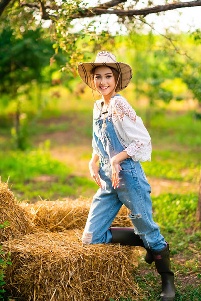 Beautiful blonde cowgirl in straw hat, checkered shirt and denim overall sitting on a straw on green nature background. Colorful art work. - Photo, Image