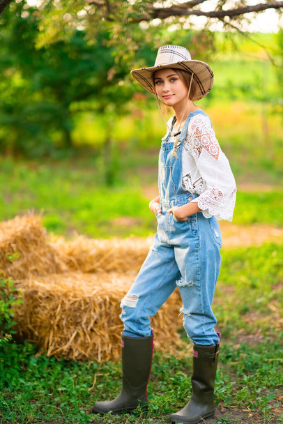 Beautiful blonde cowgirl in straw hat, checkered shirt and denim overall sitting on a straw on green nature background. Colorful art work. - Foto, Imagem