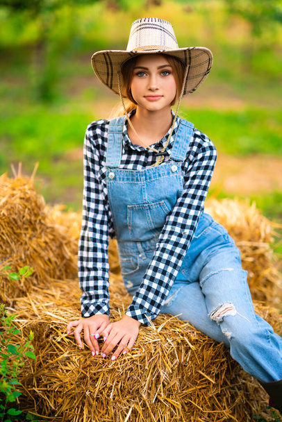 Beautiful blonde cowgirl in straw hat, checkered shirt and denim overall sitting on a straw on green nature background. Colorful art work. - Foto, Imagem