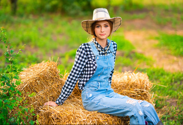 Gyönyörű szőke cowgirl szalmakalapban, kockás ing és farmer általában ül egy szívószál zöld természet háttér. Színes művészeti munka. - Fotó, kép