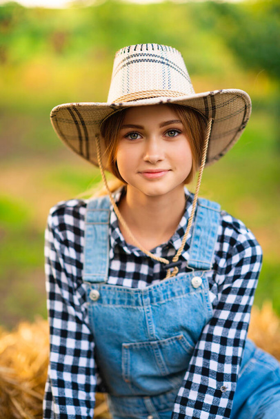 Beautiful blonde cowgirl in straw hat, checkered shirt and denim overall sitting on a straw on green nature background. Colorful art work. - Foto, Imagen