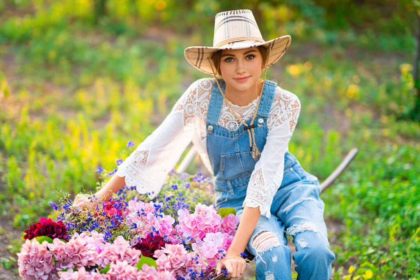 Όμορφη ξανθιά καουμπόισσα σε ψάθινο καπέλο, καρό πουκάμισο και τζιν συνολικά κάθεται σε καροτσάκι με λουλούδια στο πράσινο φόντο της φύσης. Πολύχρωμο έργο τέχνης. - Φωτογραφία, εικόνα