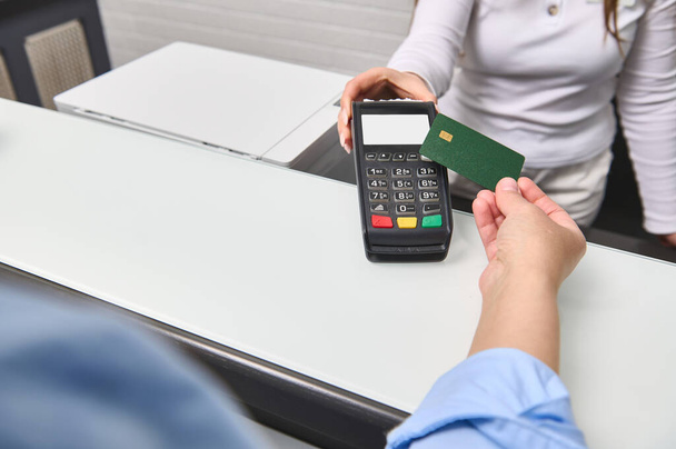 Χέρι του πελάτη πληρώνει με ανέπαφη πιστωτική κάρτα με την τεχνολογία NFC. Γυναίκα διαχειριστής με συσκευή ανάγνωσης πιστωτικών καρτών στο μετρητή υποδοχής με θηλυκό εκμετάλλευση κενή τραπεζική κάρτα. Επιλεκτική εστίαση - Φωτογραφία, εικόνα
