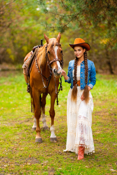 Krásná kovbojka s extra dlouhými spletené vlasy v retro krajkové šaty a oranžový klobouk pózovat na ranči s koněm. Portrét hezkého modelu na venkově. Teplé umění. - Fotografie, Obrázek