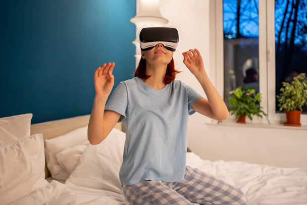 Vrouw met een virtual reality bril op bed thuis. Meisje speelt videospelletjes door middel van speciaal apparaat, helm simulator. gelukkig vrouw het dragen van speciale bril voor VR games verkent niet echt wereld - Foto, afbeelding