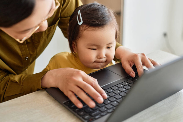 Високий кут зору Азійська мама працює онлайн на ноутбуці за столом, поки її дитина сидить на колінах. - Фото, зображення