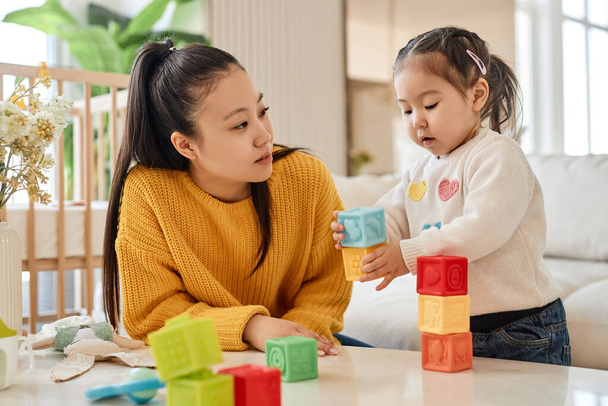 Asiatische junge Mutter spricht und spielt mit ihrer kleinen Tochter in bunten Blöcken im Zimmer - Foto, Bild