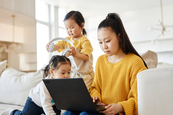 Asijské mladé maminka pracuje na notebook zatímco její děti hrát v blízkosti ní na pohovce v obývacím pokoji - Fotografie, Obrázek