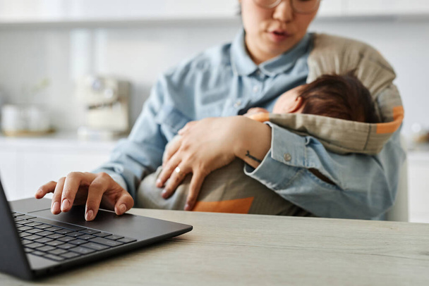 Maman tenant son bébé et travaillant en ligne sur ordinateur portable à la table dans la cuisine - Photo, image