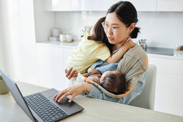 Asiatische junge Mutter mit zwei Kindern arbeitet online am Laptop, während sie in der Küche sitzt - Foto, Bild