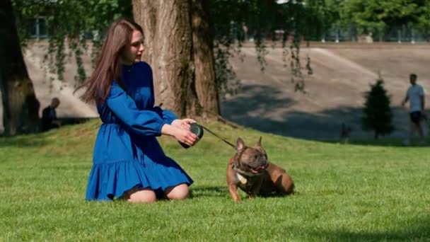uma menina bonita em um passeio com um bulldog francês no parque brinca com um cachorrinho bonito na trela - Filmagem, Vídeo