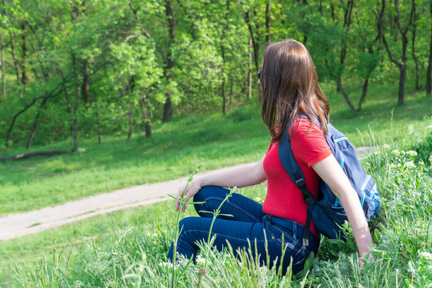 Tourist woman with backpack sit on edge and look on deciduous forest. Active adult leisure of hiking in green valley. Person enjoy wanderlust and view of the spring grassy hill. Female hiker explore. - Photo, Image