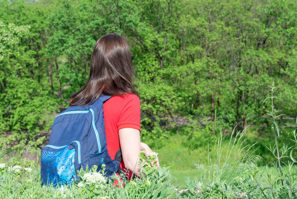 Touristin mit Rucksack sitzt am Rand und blickt auf Laubwald. Aktive Freizeit für Erwachsene beim Wandern im grünen Tal. Man genießt Fernweh und den Blick auf den frühlingshaften Grashügel. Wanderinnen erkunden. - Foto, Bild