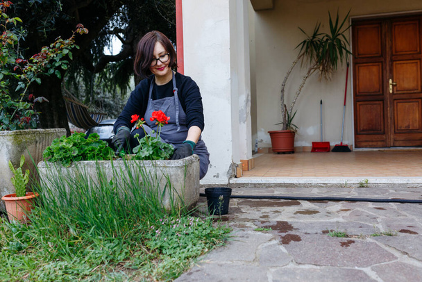 dark hair woman in apron planting floers in the pot in the garden in front of the house - Photo, Image