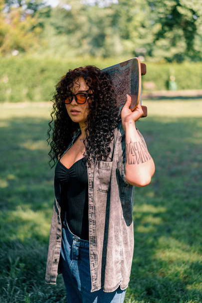 beautiful curly girl in glasses with a tattoo skater holding a skate in her hands in park resting after training summer hipsters - Фото, изображение