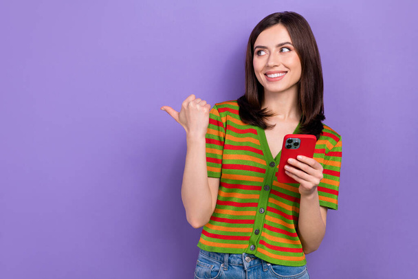 Photo of pleasant friendly nice woman wear colorful t-shirt directing look empty space hold smartphone isolated on purple background. - Фото, изображение