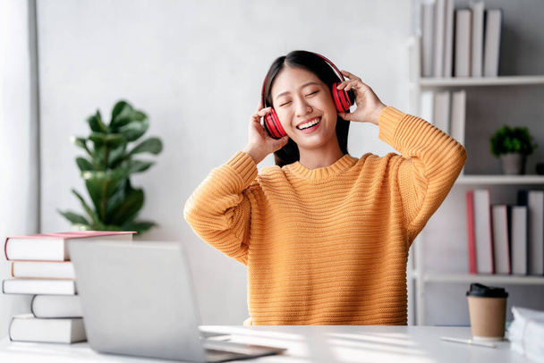 Distance education learning concept, Young woman in sweater wearing headphone and listening the music with enjoying after reading a book and studying education lesson online at home. - 写真・画像