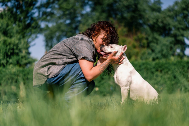 Hundepsychologe beruhigt einen weißen Hund einer großen Pitbull-Rasse im Park, ein Haustier mit herausgestreckter Zunge - Foto, Bild