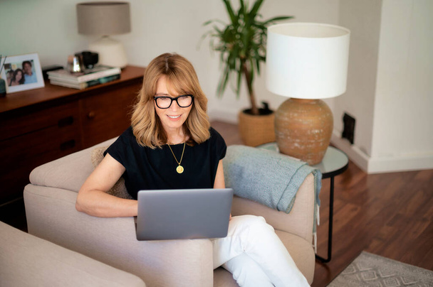 Portrait of attractive middle aged woman sitting at home and using laptop. Blond haired female wearing eyeglasses and black shirt. Home office. - Photo, Image