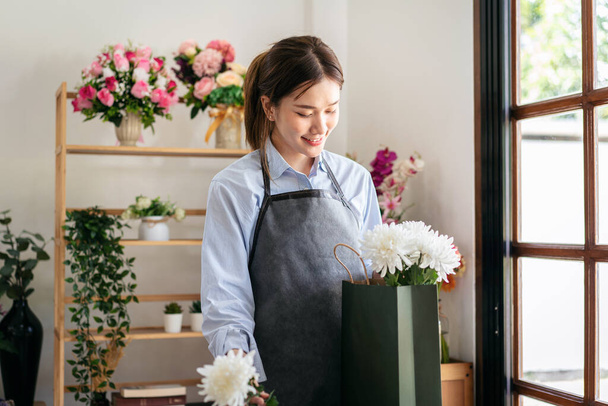 Девушка-флористка в фартуке, раскладывая белый хризантем в бумажном пакете, создавая и проектируя цветочный букет для оформления цветов в цветочном магазине. - Фото, изображение