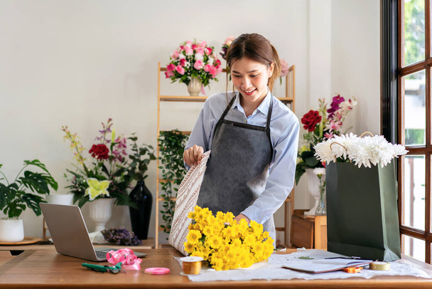 Female florist in apron using craft paper to wrapping yellow chrysanthemum on the table while creating and designing floral for arrangement flower bouquet in her flower shop. - Photo, Image
