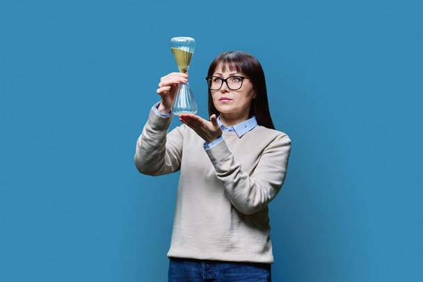 Mature woman holding an hourglass in her hands, on a blue studio background. Time, symbol past present future, passing life health beauty concept - Photo, image