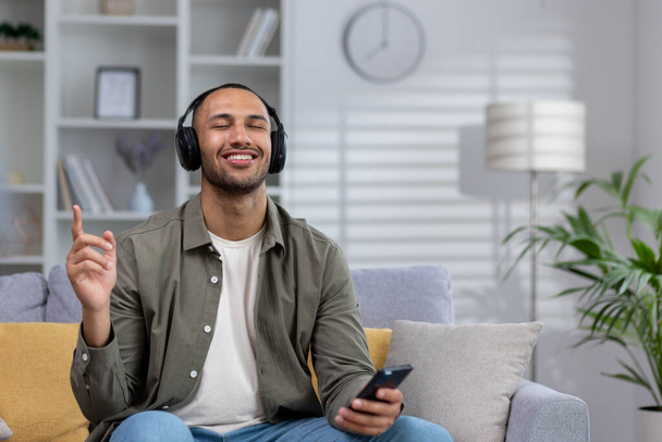 African american young man listening to music at home on sofa. He is holding a phone and wearing headphones. He closed his eyes and danced, rested, enjoyed. - Photo, Image
