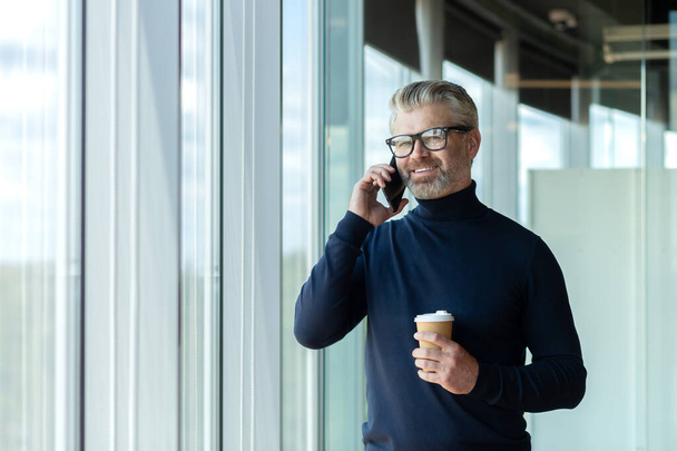 Portrait of a senior handsome gray-haired man designer, architect, engineer standing in the office by the window talking on the phone and holding a cup of coffee. Smiling looking at the camera. - 写真・画像